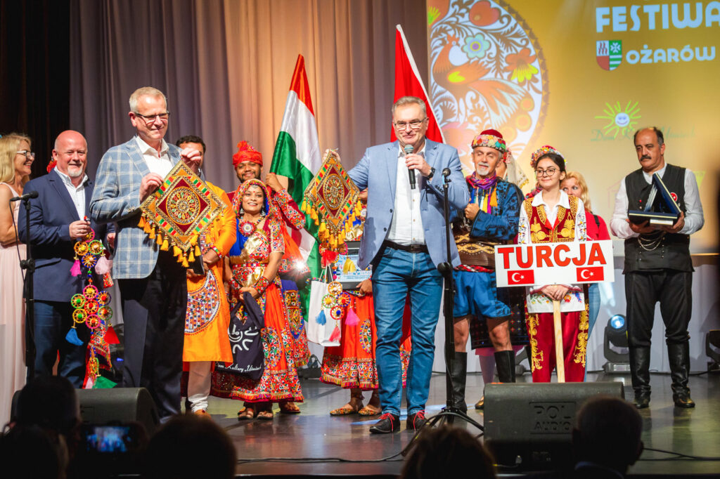 Na zdjęciu widoczni są organizatorzy wydarzenia, stoją na scenie, w tle członkowie zespołów uczestniczących w Międzynarodowym Festiwalu Folkloru