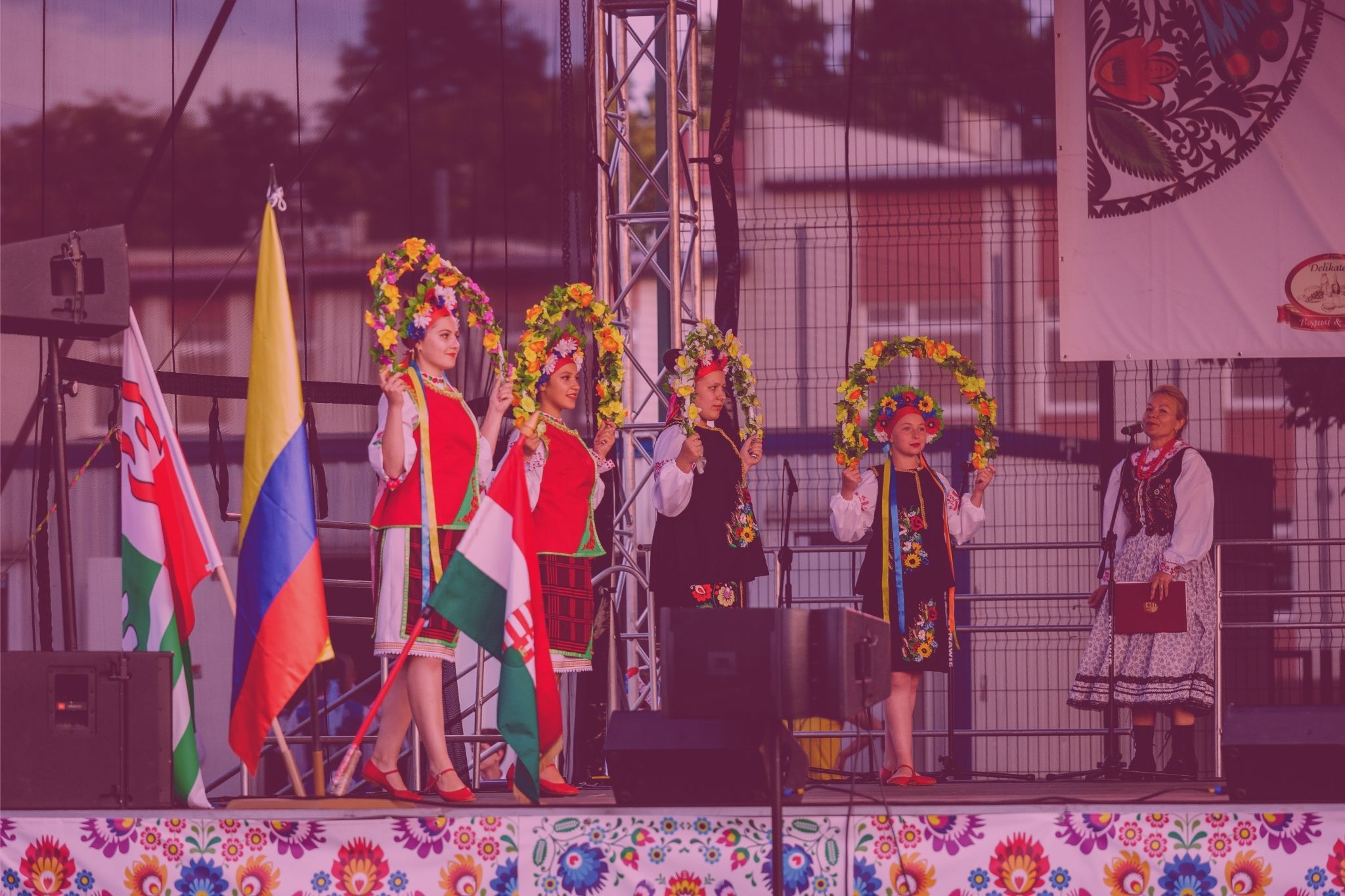 Zdjęcie przedstawia pięć kobiet w strojach ludowych, jedna z nich ma polski strój ludowy i stoi przy mikrofonie, cztery kobiety występują na scenie